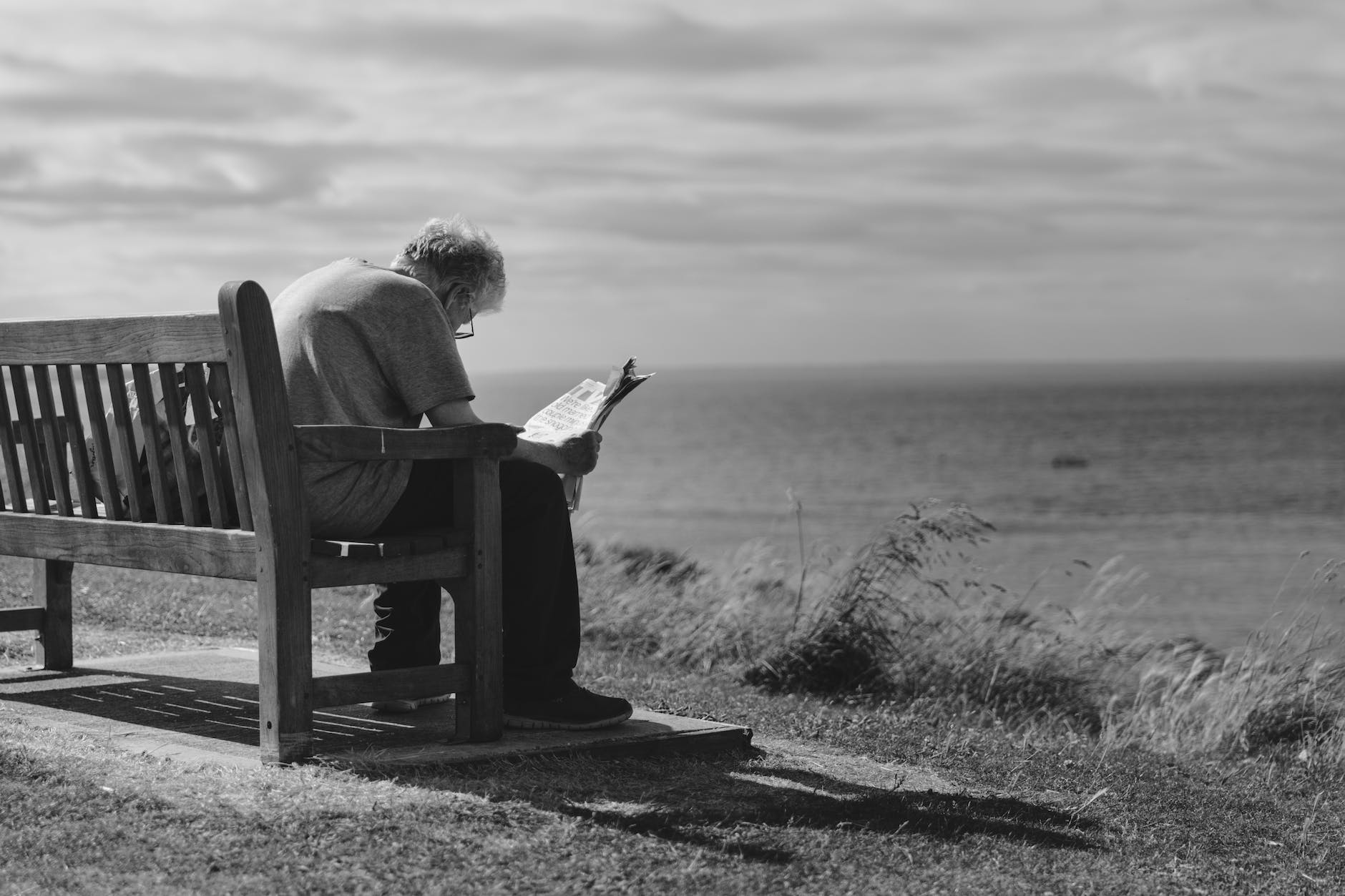 grayscale photo of man sitting on brown wooden bench reading news paper during day time